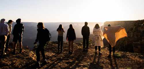 A group of students stands at the edge of a cliff viewing the sunrise. The sunlight catches on the blankets they have wrapped around themselves to keep warm.